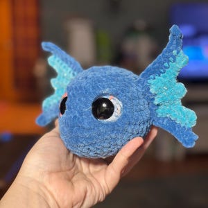 May include: A round, blue, crocheted plush toy featuring large, glossy black eyes and two prominent, textured, light blue and dark blue fin-like appendages extending from the sides of its head, crafted from soft, fuzzy yarn material.