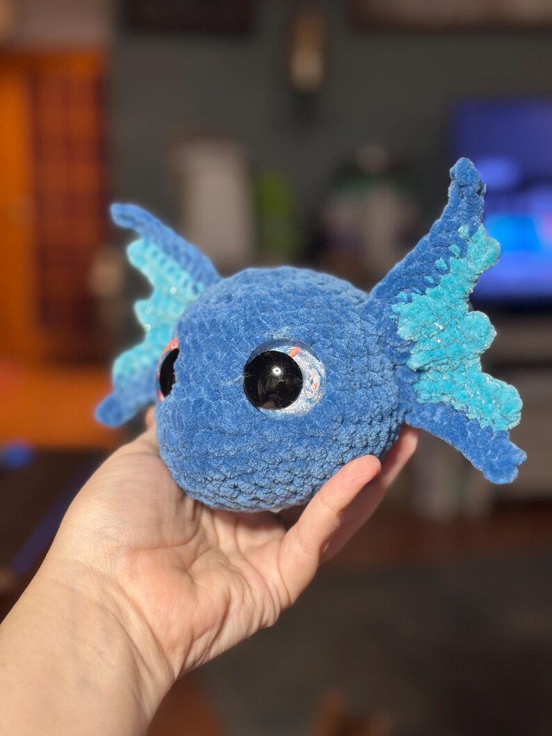 May include: A round, blue, crocheted plush toy featuring large, glossy black eyes and two prominent, textured, light blue and dark blue fin-like appendages extending from the sides of its head, crafted from soft, fuzzy yarn material.