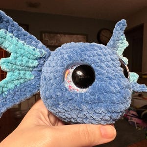 May include: A plush toy crafted from blue and light blue yarn, featuring a rounded head and large, dark, glossy eyes with a reflective surface. The creature has prominent, textured side fins in alternating shades of blue and light blue.