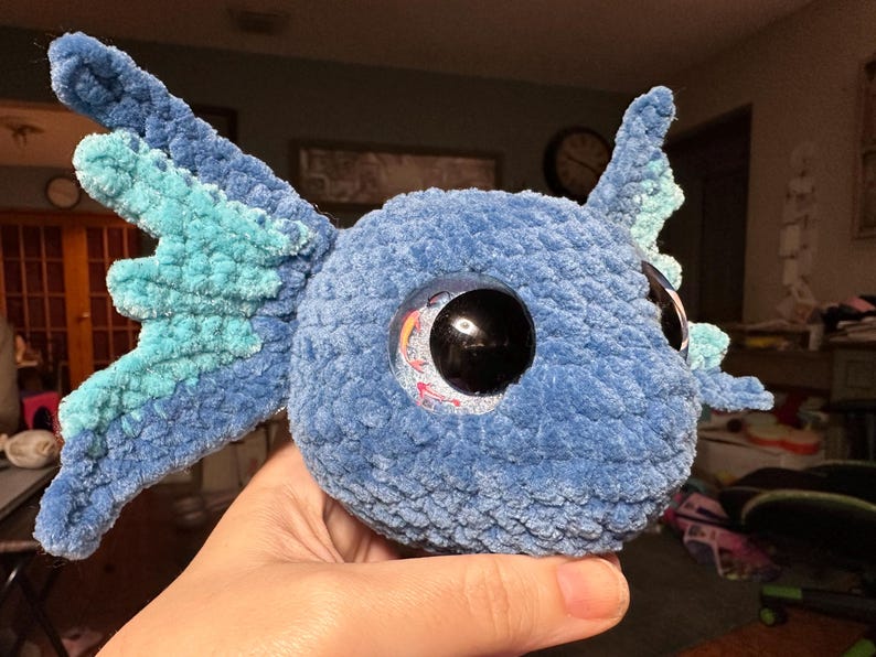 May include: A plush toy crafted from blue and light blue yarn, featuring a rounded head and large, dark, glossy eyes with a reflective surface. The creature has prominent, textured side fins in alternating shades of blue and light blue.