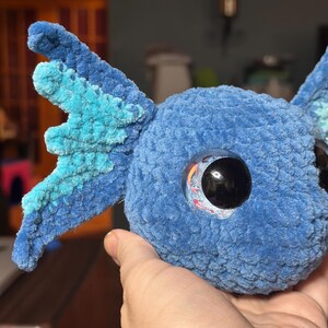 May include: A blue crocheted plush toy with a rounded head and large, textured side fins in shades of light and dark blue. The toy features a prominent, glossy black eye with a small, colourful koi fish graphic visible inside the iris area.