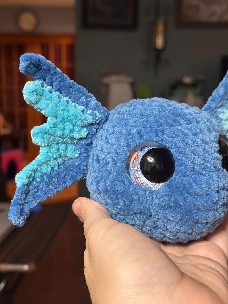 May include: A blue crocheted plush toy with a rounded head and large, textured side fins in shades of light and dark blue. The toy features a prominent, glossy black eye with a small, colourful koi fish graphic visible inside the iris area.