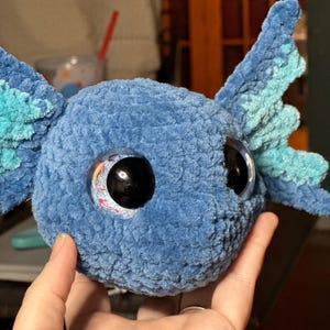 May include: A blue crocheted plush toy shaped like an axolotl head. The plush features a soft, textured yarn surface, large black plastic eyes with a decorative koi fish graphic inside, and prominent, light blue, fan-shaped external gills.