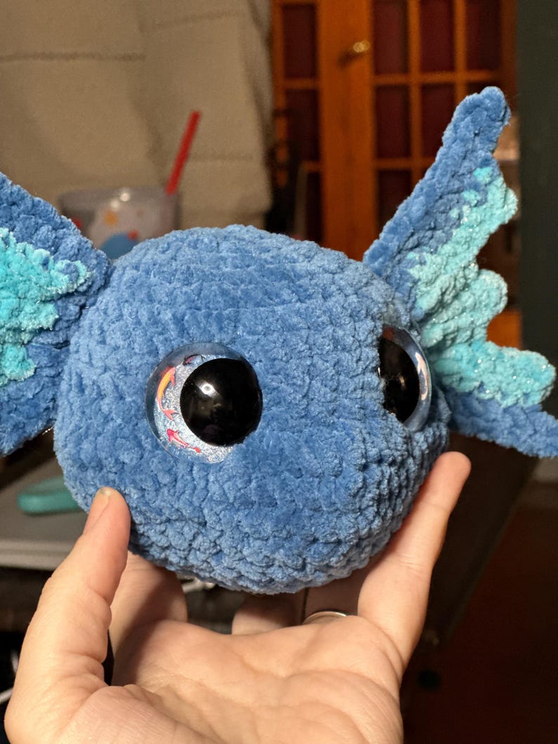 May include: A blue crocheted plush toy shaped like an axolotl head. The plush features a soft, textured yarn surface, large black plastic eyes with a decorative koi fish graphic inside, and prominent, light blue, fan-shaped external gills.
