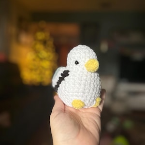 May include: A white crocheted bird with black and yellow accents. The bird is sitting in a person's hand.