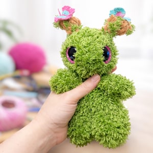 May include: A green plush creature with large, glittery eyes and small butterfly accents on its ears. The handmade toy has a fuzzy texture and is held in a person's hand. The background includes colorful yarn balls.