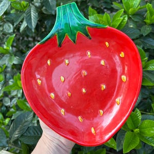 Op de afbeelding: Een rode, keramische aardbeivormige serveerschaal met een groene steel. De schaal heeft gele zaaddetails en wordt tegen een achtergrond van groen gebladerte gehouden. Deze decoratieve schaal is ideaal voor het serveren van snacks of het presenteren van fruit.