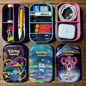 May include: A selection of small, colourful storage containers and Pokémon trading card tins. The containers hold items such as scissors, thread, batteries, and charging cables. The card tins feature Pokémon characters and the text "Pokémon Trading Card Game."