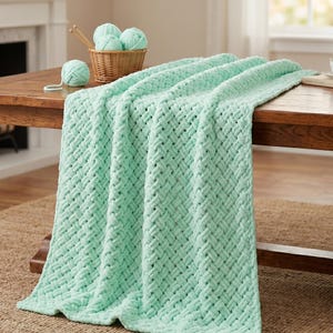 May include: A mint green woven blanket draped over a wooden table. The blanket has a thick, textured weave. A small basket with several balls of yarn and knitting needles sits on the table. A single ball of yarn rests nearby.