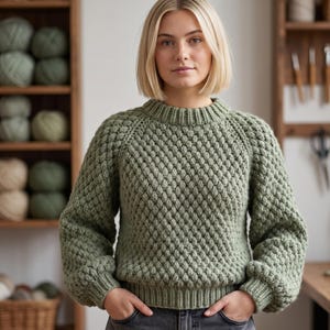 Puede incluir: Un suéter tejido a mano de color verde salvia con un patrón texturizado en forma de diamante. El suéter tiene cuello redondo y mangas largas con puños acanalados. La persona lleva el suéter con las manos en los bolsillos.