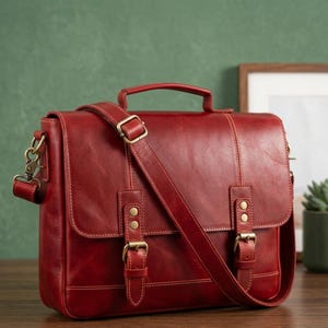 Op de afbeelding: Een rode leren tas met een handvat aan de bovenkant en een verstelbare schouderriem. De tas heeft messingkleurige gespen en hardware. De tas heeft een klepsluiting en is ontworpen met zichtbare stiksels.