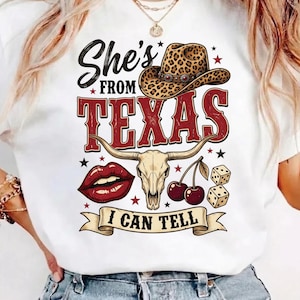 Może przedstawiać: Biała koszulka z grafiką z napisem "She's From Texas" w kolorze czarnym i czerwonym, kowbojskim kapeluszem w panterkę, czaszką byka, wiśniami, kostkami do gry i czerwonymi ustami. Na banerze widnieje napis "I Can Tell".