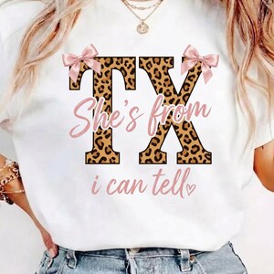 May include: White t-shirt featuring a leopard print "TX" design, pink bows, and the text "She's from TX i can tell" in pink script. A casual style top.