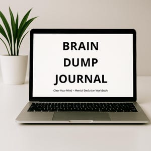 Puede incluir: Un ordenador portátil plateado muestra el texto "BRAIN DUMP JOURNAL" en negro. Debajo, un texto más pequeño dice "Clear Your Mind - Mental Declutter Workbook". Una planta en maceta es visible en el fondo.
