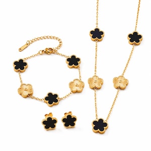 Puede incluir: Un conjunto de joyas doradas que incluye una pulsera, un collar y pendientes. Cada pieza presenta un diseño en forma de trébol con centros negros y bordes dorados texturizados. El conjunto se muestra sobre un fondo blanco.