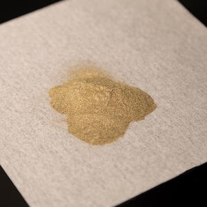 Könnte beinhalten: Ein kleiner Haufen feines, goldfarbenes Pulver liegt auf einer hellgrauen, strukturierten Oberfläche. Das Pulver scheint sich in der Mitte zu konzentrieren, mit einer leicht ungleichmäßigen Verteilung. Der Hintergrund ist neutral.