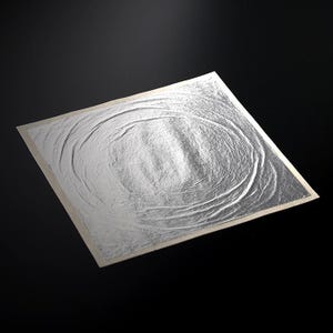 Puede incluir: Una lámina cuadrada de pan de plata con una superficie texturizada y un patrón circular. La hoja de plata tiene un brillo metálico y está colocada sobre un fondo oscuro. Los bordes de la hoja son de color beige claro.