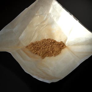 Pode incluir: Uma pequena pilha de pó castanho claro repousa sobre um papel dobrado, esbranquiçado. O papel está aberto, revelando o pó, contra um fundo escuro. O pó parece ser finamente moído.