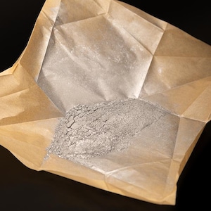 May include: A pile of gray powder sits in a partially opened, folded brown paper packet. The powder has a textured, granular appearance. The paper is creased and has a matte finish, with a dark background.