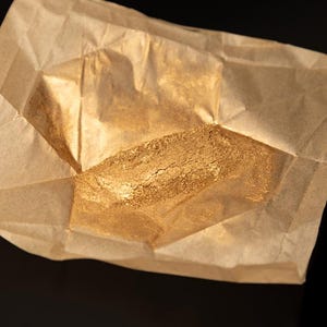 Può includere: Primo piano di polvere d'oro in un pacchetto di carta marrone. La polvere ha un aspetto strutturato e una lucentezza metallica. La carta è piegata e spiegazzata, con pieghe e ombre visibili. Lo sfondo è nero.