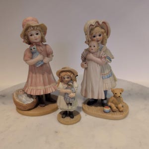 Könnte beinhalten: Eine Sammlung von Porzellanfiguren, die junge Mädchen darstellen. Eines hält eine blaue Katze, ein anderes eine Puppe und ein drittes ein Baby. Ein kleiner Teddybär und ein Ball sind ebenfalls vorhanden. Die Figuren sind in Pastellfarben gehalten und haben detaillierte Merkmale.