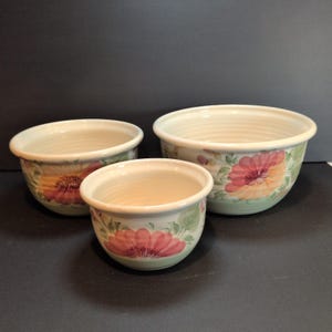 May include: A set of three ceramic bowls of varying sizes, each featuring a cream-colored interior and a painted exterior design of pink and orange flowers with green leaves. The bowls have a rounded shape with a smooth, glossy finish throughout.