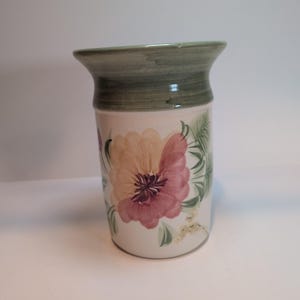 May include: A cylindrical ceramic vase with a flared rim, featuring a hand-painted floral design in shades of pink, peach, and green on a cream-colored background. The upper portion of the vase is finished with a solid, muted green band.
