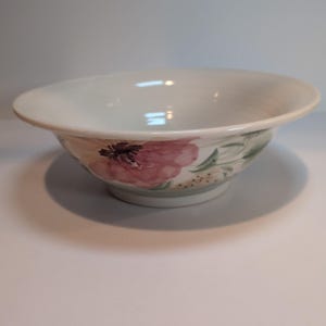 May include: A white ceramic bowl with a wide, flared rim and a rounded base. The exterior features a painted floral design with large pink petals, dark centers, and green foliage accents. The interior of the bowl is plain white and smooth.