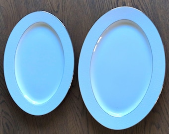 May include: Two oval-shaped white ceramic serving platters of different sizes. Each platter features a smooth, solid white center basin surrounded by a wide, textured white rim with a thin, metallic silver border along the outer edge.