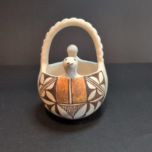 Puede incluir: Una pequeña cesta de cerámica blanquecina con asa. La cesta presenta una figura de pájaro y está decorada con patrones geométricos marrones y blancos. El centro tiene un diseño naranja y marrón.