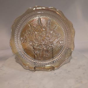 May include: A clear, iridescent glass plate with a scalloped edge. The plate features a detailed embossed design of a floral bouquet in the center. The plate is displayed on a metal stand, showcasing its decorative and vintage appeal.