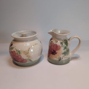 May include: A ceramic sugar bowl with a lid and a matching creamer pitcher. Both pieces feature a cream-colored base with a painted floral pattern in shades of pink, peach, and green, finished with a soft green band around the bottom edge.