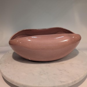 May include: A pink ceramic bowl with a speckled texture. The bowl has a unique, organic shape with gently curved sides. The interior is a darker shade of pink, and the bowl sits on a white marble surface.