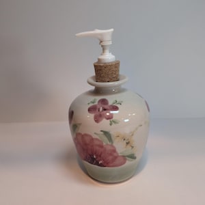 May include: A ceramic soap dispenser with a rounded body featuring hand-painted floral motifs in shades of deep red, pink, and yellow. The dispenser includes a natural cork stopper fitted into the neck and a white plastic pump mechanism on top.