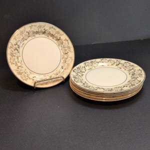 Puede incluir: Un juego de platos redondos de cerámica con un centro de color crema y un borde decorativo con un patrón floral en dorado metálico. Un plato se muestra en posición vertical sobre un soporte, mientras que una pila de cuatro platos iguales descansa al lado.