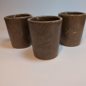 Może przedstawiać: Trzy brązowe kubki ceramiczne z nakrapianym wykończeniem i gładkim, zwężającym się cylindrycznym kształtem. Każdy kubek posiada małe, wytłoczone logo na przedniej powierzchni, przedstawiające stylizowanego ptaka w locie. Ziemiste odcienie.