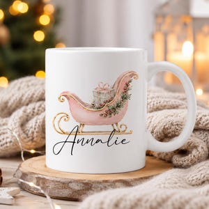 May include: A white ceramic mug featuring a pink sleigh illustration with gold runners, a gift box, and holly accents. The name Annalie is printed in black cursive script below the sleigh. The mug has a standard handle and a smooth finish.