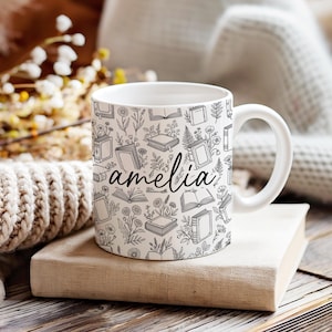 May include: White ceramic mug featuring a black and white book and floral design. The name "amelia" is written in cursive on the front. The mug is placed on a stack of books.