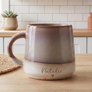 May include: A ceramic mug featuring a two-tone gradient design that transitions from a dark brown at the top to a light cream colour at the base. The name Natalie is printed in a dark script font above a small heart icon on the lower section.