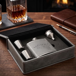 May include: A grey gift box containing a stainless steel flask engraved with the name HENRY, two matching stainless steel shot cups, and a small stainless steel funnel. The interior of the box is lined with dark grey felt to hold each item.