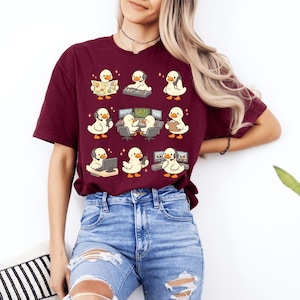 May include: Burgundy t-shirt featuring a grid of cartoon ducks in various work-related scenarios. The ducks are depicted with headsets, computers, and maps. The design is playful and whimsical, perfect for those who enjoy a touch of humor in their wardrobe.