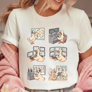 May include: White t-shirt featuring a cartoon duck in various scenes, including using computers and a map. The duck is depicted in different poses, suggesting a theme of technology and problem-solving. The design is playful and whimsical.
