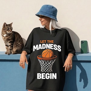 May include: Black t-shirt with the text "LET THE MADNESS BEGIN" and a basketball graphic. A woman wearing a denim bucket hat is leaning against a blue surface. A tabby cat sits to the left of the woman. A green phone is on the right.