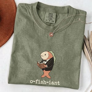 以下が含まれることがあります： 黒いタキシードと蝶ネクタイを身につけ、開いた本を持っている魚のグラフィックが描かれたオリーブグリーンのTシャツ。魚の下には「o-fish-iant」という文字があります。Tシャツはクルーネックで、Comfort Colorsのブランドラベルが付いています。