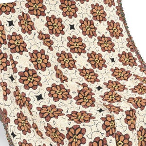 May include: A woven throw blanket with a repeating floral pattern in shades of brown, tan, and black. The blanket is draped over a brown bench, showcasing its fringed edges and intricate design. The throw blanket is a decorative home textile.