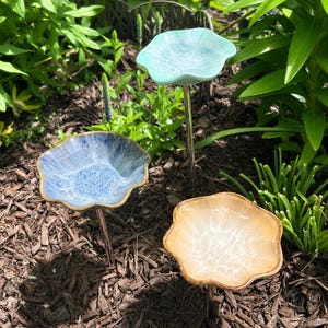 May include: Three garden stakes featuring flower-shaped bowls in light blue, dark blue, and tan. Each bowl has a wavy, scalloped edge and a glossy finish, mounted on a thin metal rod. The stakes are positioned at varying heights in the soil.