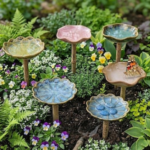 Könnte beinhalten: Eine Sammlung von Vogeltränken aus Keramik in verschiedenen Farben, darunter Grün, Rosa und Blau, mit einem gewellten Rand. Jede Tränke wird von einem Holzpfosten getragen und befindet sich in einem Garten mit Blumen und Grün. Ein Schmetterling sitzt auf einer der Tränken.