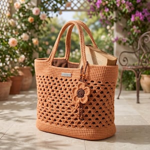 Peut inclure: Un sac cabas en crochet couleur pêche avec un motif ajouré en losanges, deux anses solides et une base pleine. Une petite breloque fleur en crochet marron foncé avec des perles en bois pend à l'anse, et un livre est glissé à l'intérieur.