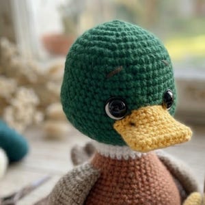 Puede incluir: Un pato de ganchillo hecho a mano con cabeza verde, pico amarillo y ojos de bot&oacute;n negros. El pato tiene un cuerpo marr&oacute;n y un cuello blanco. El pato es un juguete blando.