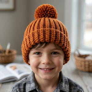 Puede incluir: Un gorro de punto acanalado de color marrón anaranjado con un gran pompón en la parte superior. El gorro está hecho de un hilo grueso y tiene un estilo clásico y cálido. El gorro lo lleva una persona.